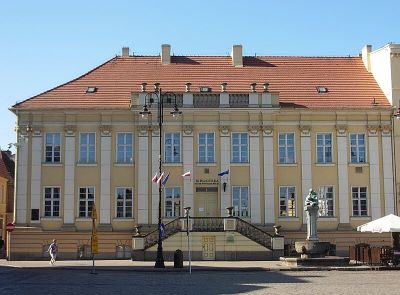 A historical building with a yellow facade and a red roof, featuring several windows and flags. A city square is visible in front of the building.